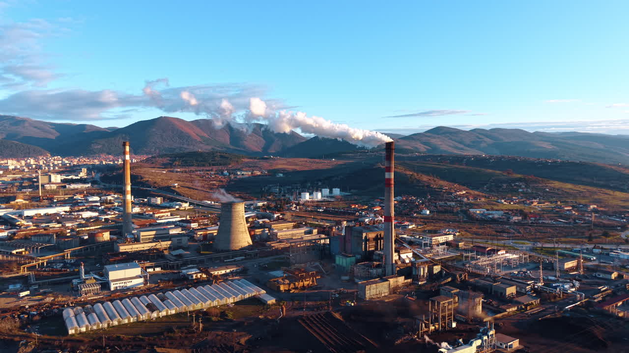 Aerial view of industrial power plant with smoking chimneys. Industrial zone with cooling towers and tall chimneys emitting smoke during sunset against mountain backdrop