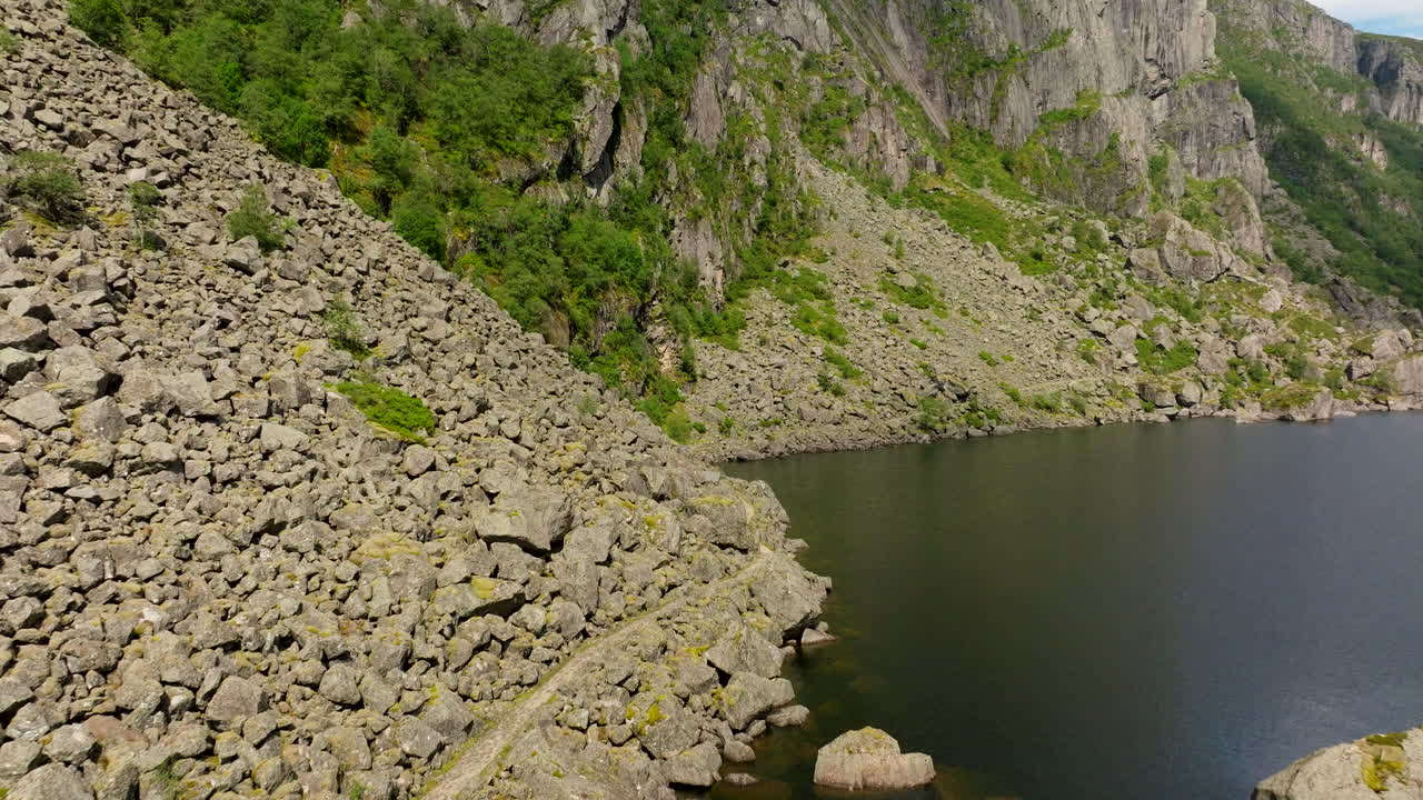 Drone advances along rocky fjord shoreline beneath steep cliffs. Aerial