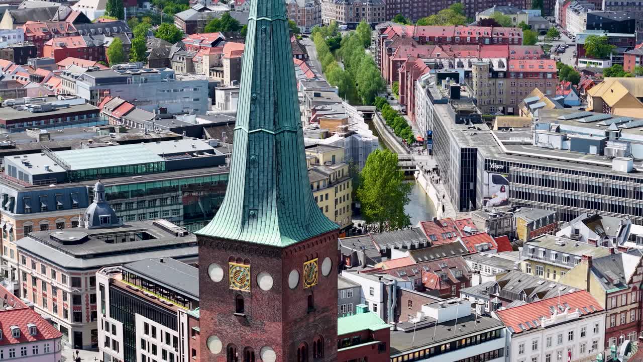 Aerial close‑up of Aarhus city center highlighting a historic church tower with a green spire, clock faces and surrounding colorful rooftops