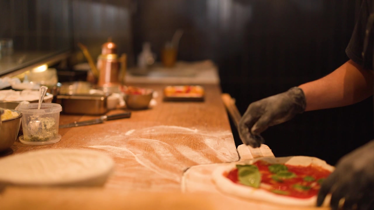 Primo piano di uno chef con guanti neri che stende l'impasto della pizza con salsa di pomodoro e basilico.