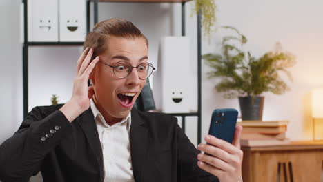 happy amazed young caucasian businessman working on smartphone shocked by sudden victory at home