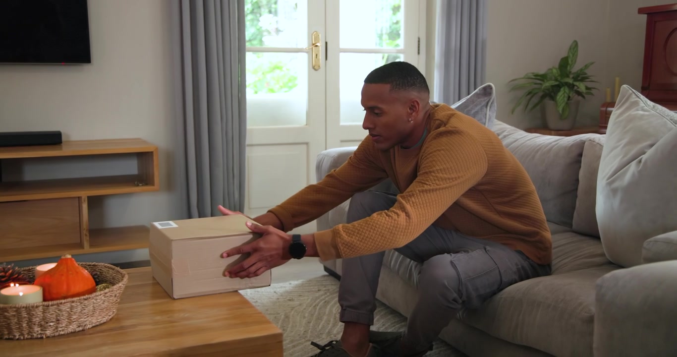 African American man on sofa checking phone, placing box on table beside woven tray, copy space
