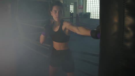 Slow-Motion-shot:-Strong-athletic-female-kickboxer-exercising-with-a-boxing-bag-in-dark-gym-with-smoke-and-kicking-a-boxing-bag