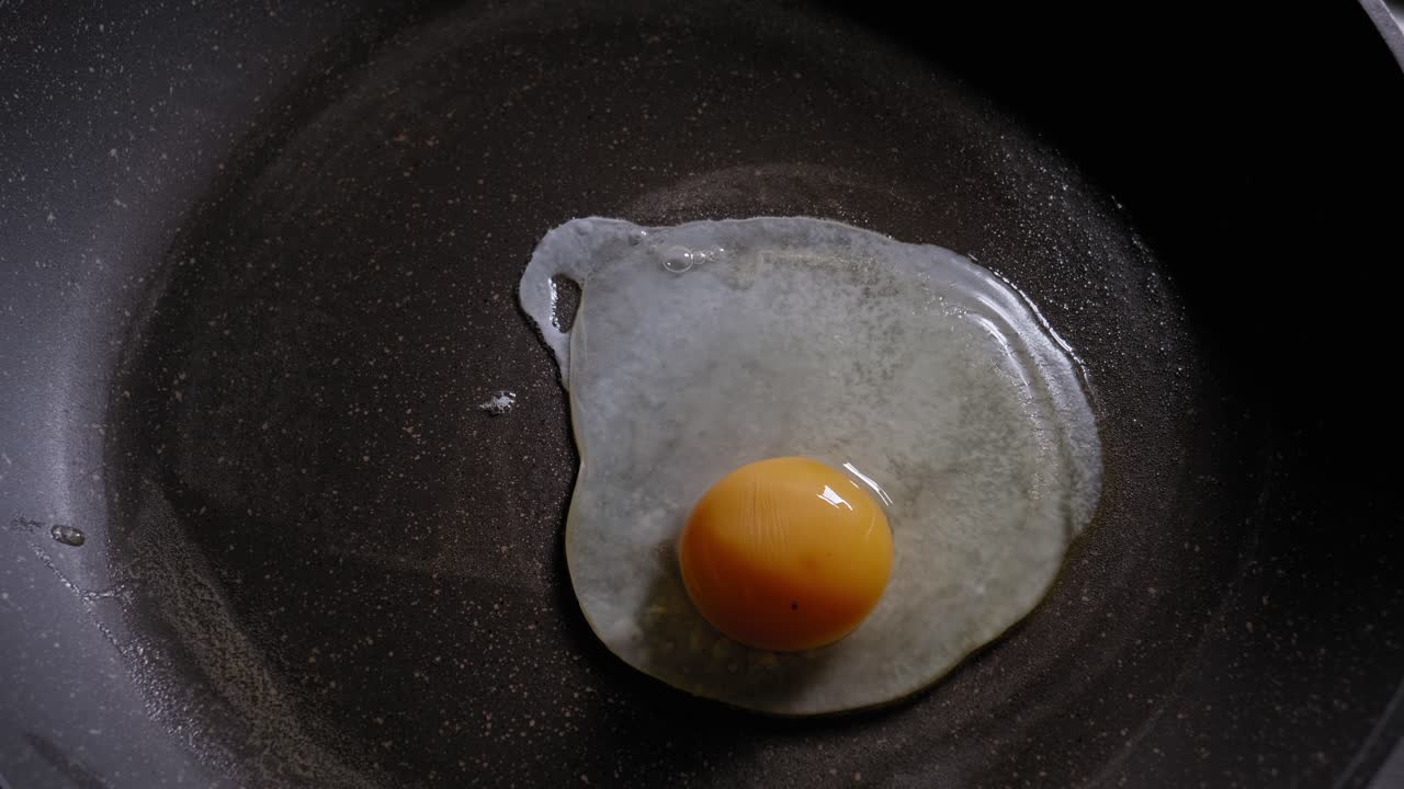 A top down close up shot of a fresh egg being poured onto a lightly greased frying pan, the raw egg changes colour as it starts to cook and splutters and bubbles on the surface of the hot pan