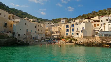 view of levanzo sicilian fishing village and waterfront scenic bay with people bathing and fishermen boats in background , sicily in italy