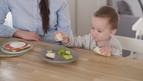 lindo bebé tomando plátano del plato y comiendo mientras su madre se sienta a su lado en casa