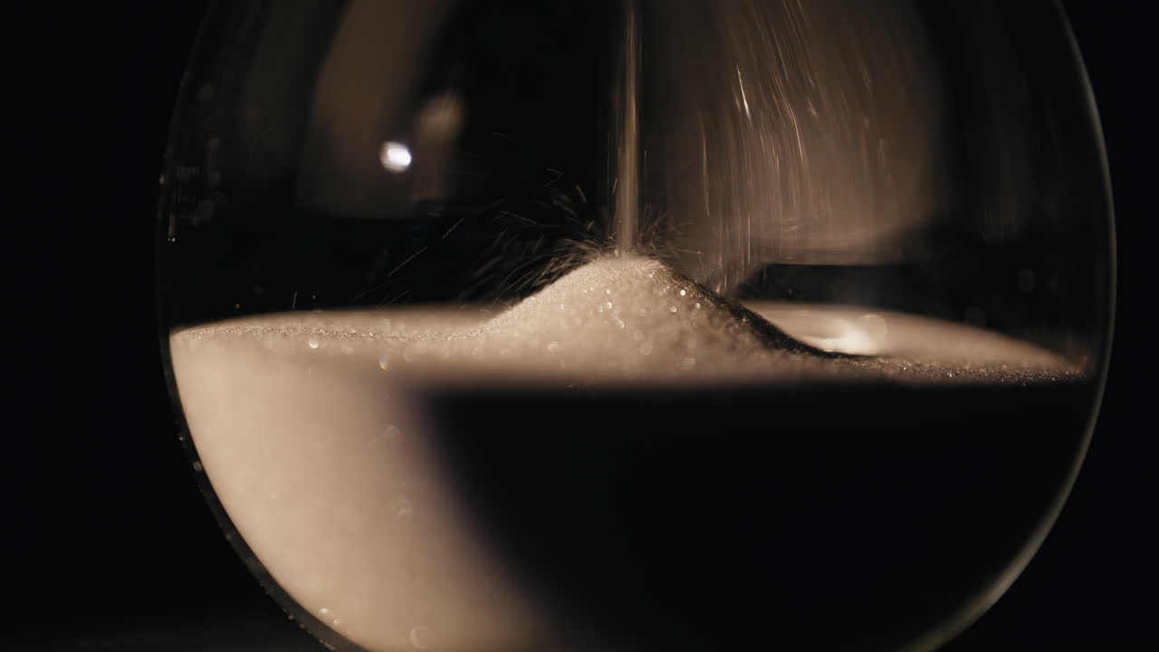 Closeup sand falling hourglass standing dark room. Gold grains flowing inside
