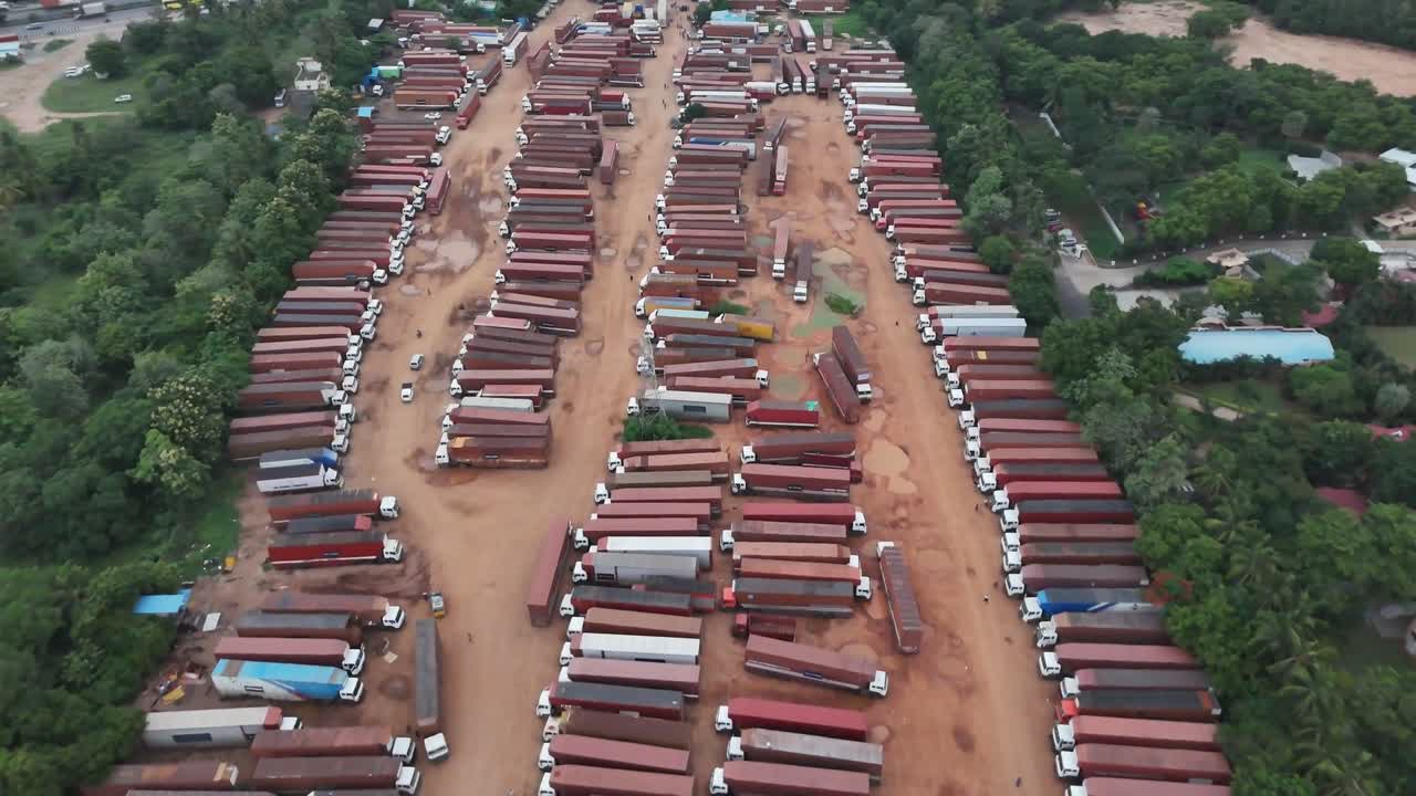 An aerial drone view of a massive fleet of commercial trucks parked in neat rows in a large depot. The truck yard is surrounded by a lush green scale of the logistics and transportation industry