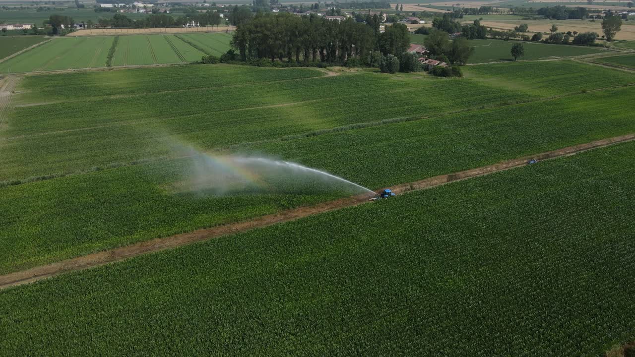 Ripresa aerea 4K con drone che ruota in senso antiorario attorno a un trattore che irriga un terreno agricolo nella Val Padana, con lo spruzzo dell'acqua che forma un arcobaleno sopra il raccolto