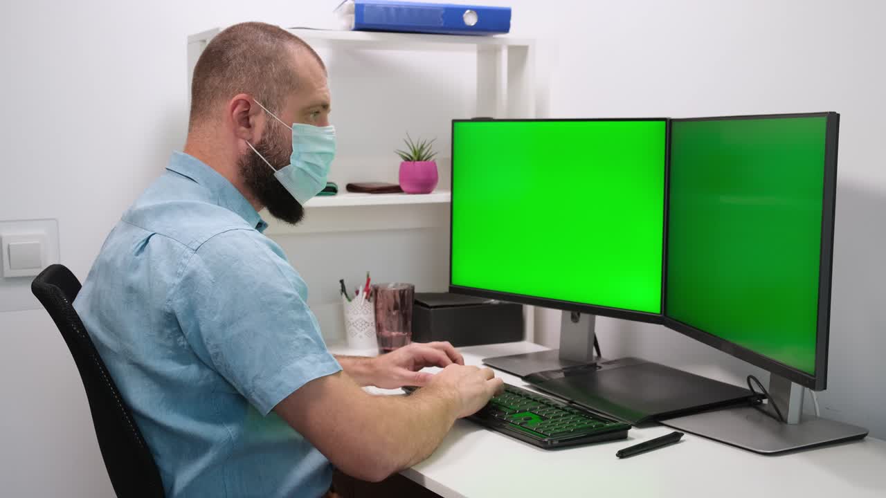 businessman freelancer working on computer at home. Two Green Screens. A man wearing a medical mask works in front of two green monitors. Remote work at home in a self-isolation quarantine