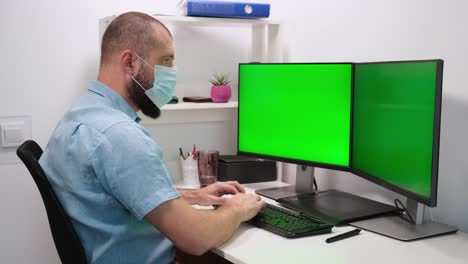 businessman freelancer working on computer at home. two green screens. a man wearing a medical mask works in front of two green monitors. remote work at home in a self-isolation quarantine