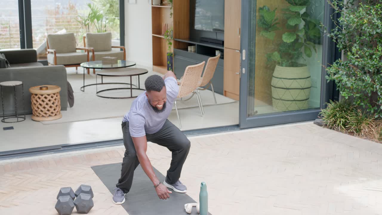 African American man in fitted tee placing feet, squatting and warming on patio mat with dumbbells