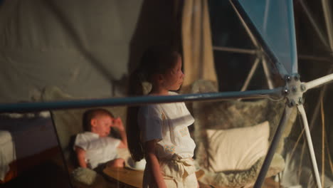 upset little girl and sleepy brother in glamping hut. children tired after active tourist day rest at luxury camp. kids wait for parents in hotel room