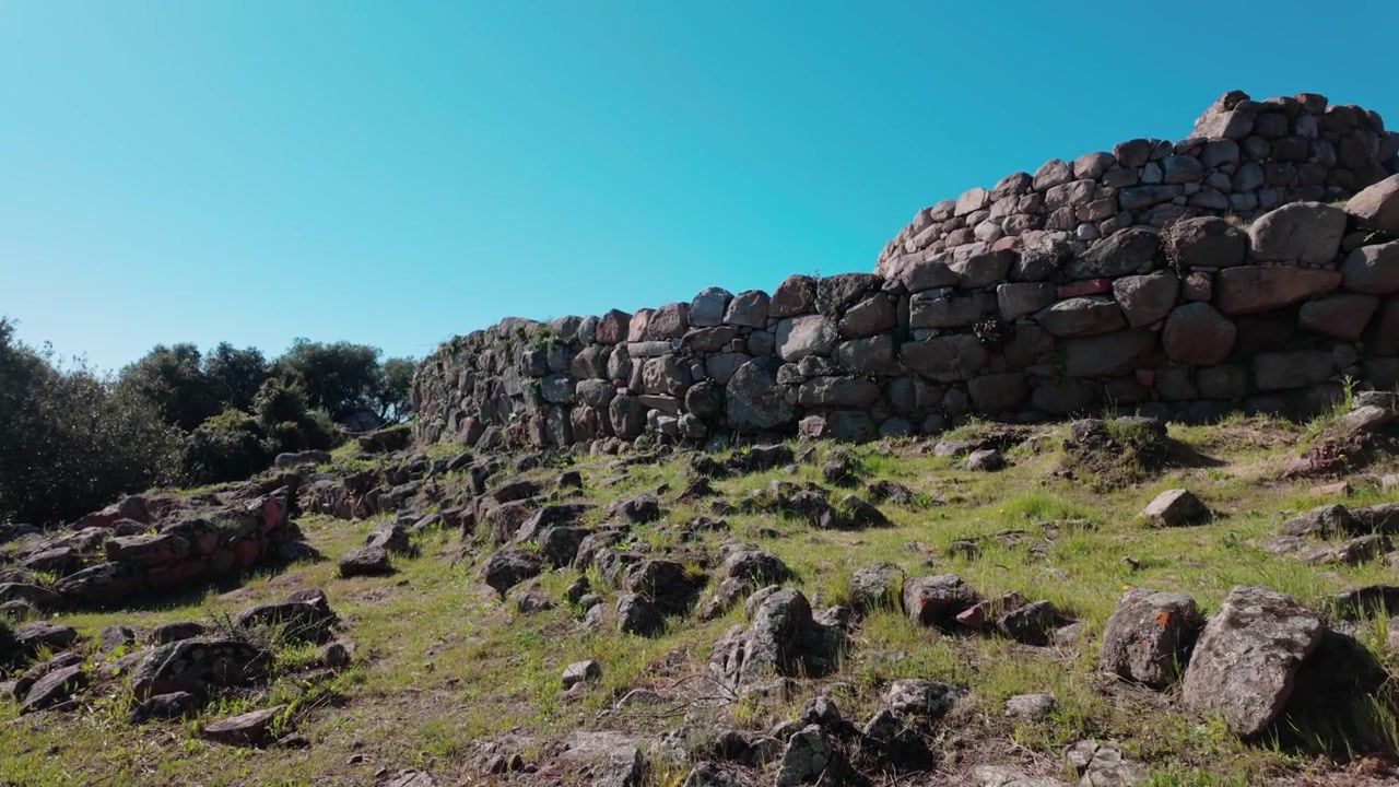 Široký záběr prehistorické archeologické lokality Nuraghe SOrtali e su Monti na Sardinii v Itálii, s prastarými suchými kamennými zdmi pod jasně modrou oblohou.
