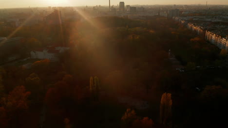 Bosque-De-árboles-De-Color-Naranja-Y-Rojo-O-Parque-Público-En-La-Gran-Ciudad,-Contraste-Entre-El-Entorno-Urbano-Y-La-Naturaleza,-La-Plataforma-Aérea-Se-Inclina-Hacia-Arriba-Sobre-Berlín,-El-Paisaje-Urbano-De-Alemania