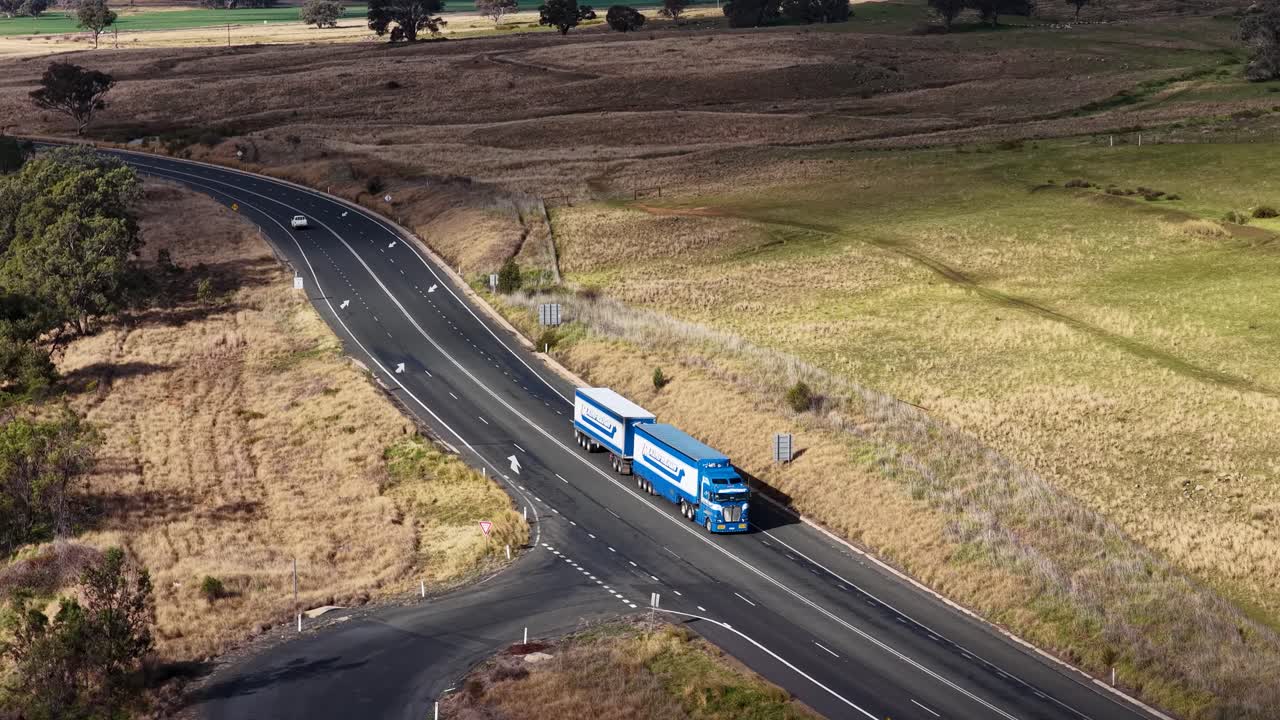A blue semi truck with trailer travels along a winding rural road in dry countryside, captured in daylight from a high, aerial perspective