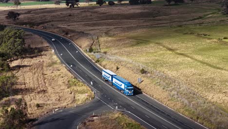 a blue semi truck with trailer travels along a winding rural road in dry countryside, captured in daylight from a high, aerial perspective