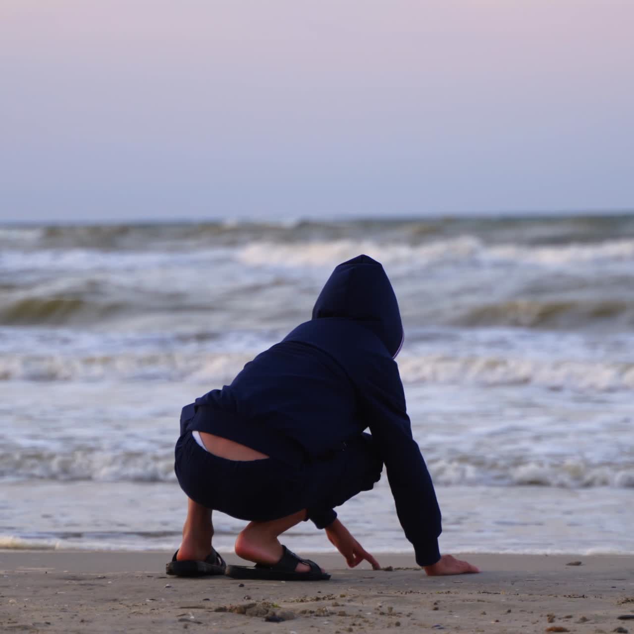 Junge ruht sich bei Sonnenuntergang am Meer aus. Das Kind malt am Sandstrand vor dem Hintergrund schäumender Wellen des Abendmeeres.