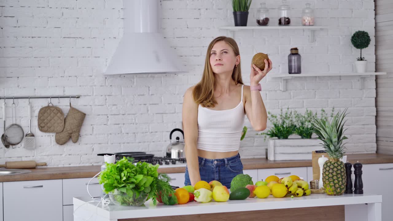 Beautiful woman with coconut after shopping. Young lady holding a fresh fruit and smiling on the kitchen background. Fresh fruits and vegetables on the table. Healthy food concept.