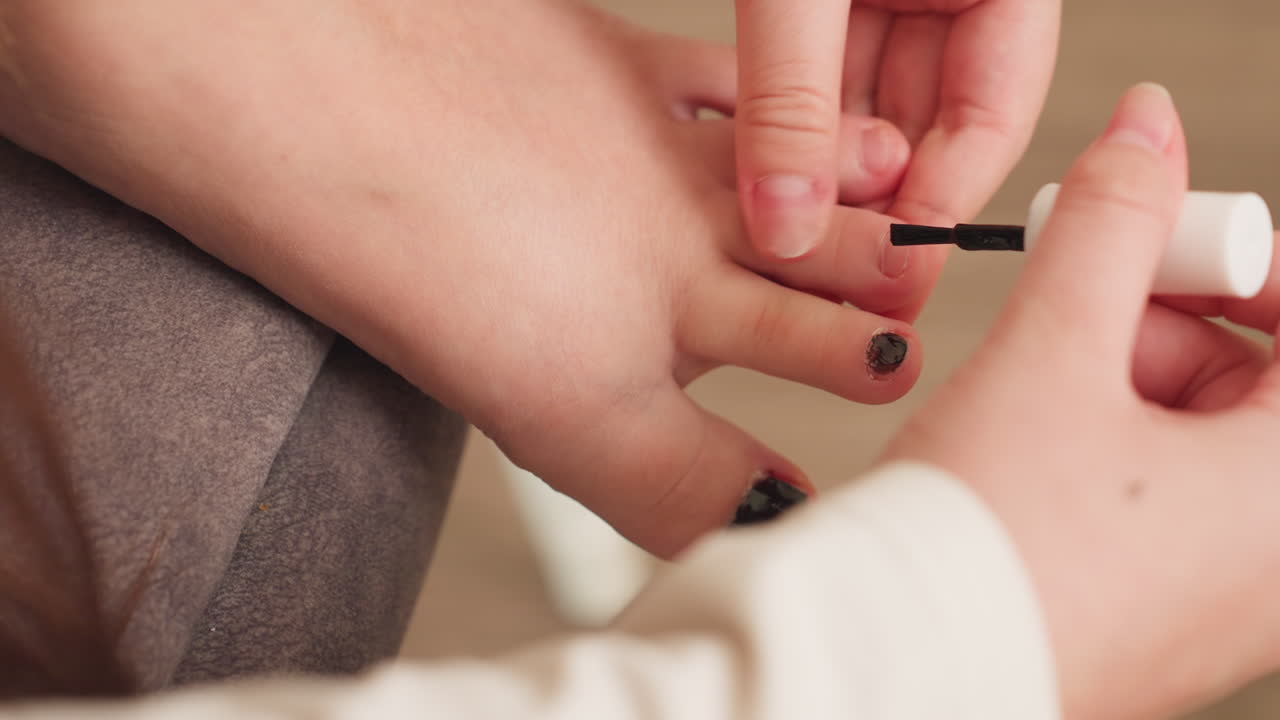 High angle view of beautician hands painting toes with black polish, close focus on careful strokes, big toe coated, relaxed home pedicure on chair cushion, soft light, tidy grooming routine