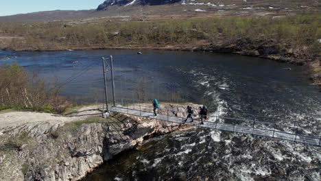 three hikers walking over a suspension bridge on the kungsleden near abiskojaure in the swedish mountains, backpacking in the forest, pan from right ot left