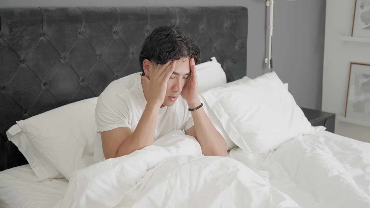 Pria dewasa berbaju kaus putih terbangun di tempat tidur karena alarm ponsel, meraih ponsel, duduk, memijat pelipis
