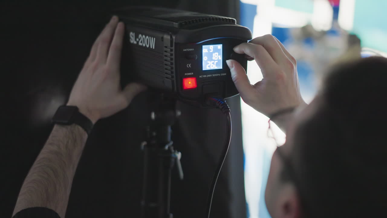 Technician adjusting power button and brightness dial on SL-200W LED studio light mounted on tripod bracket amid blue-lit studio backdrop during broadcast production setup with cables visible