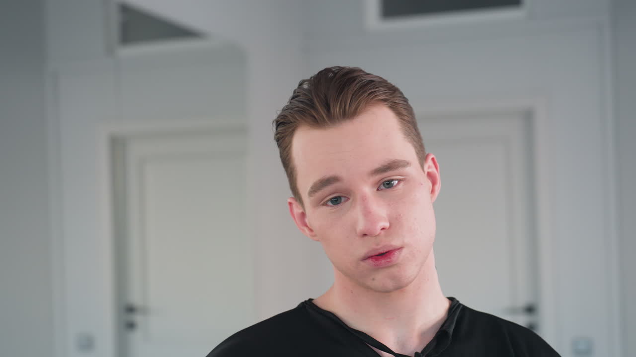 young man in black outfit standing with relaxed posture and parted lips as he takes deep breath, expressing calmness and emotional release in minimalistic interior with bright natural lighting