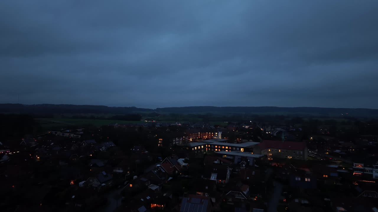 Letecký pohled na americké město za zimního soumraku, s blednoucím denním světlem nad tmavými střechami, roztroušenými teplými světly a klidným horizontem. Tichá, chladná večerní atmosféra. Osvětlená okna školní budovy. Široký