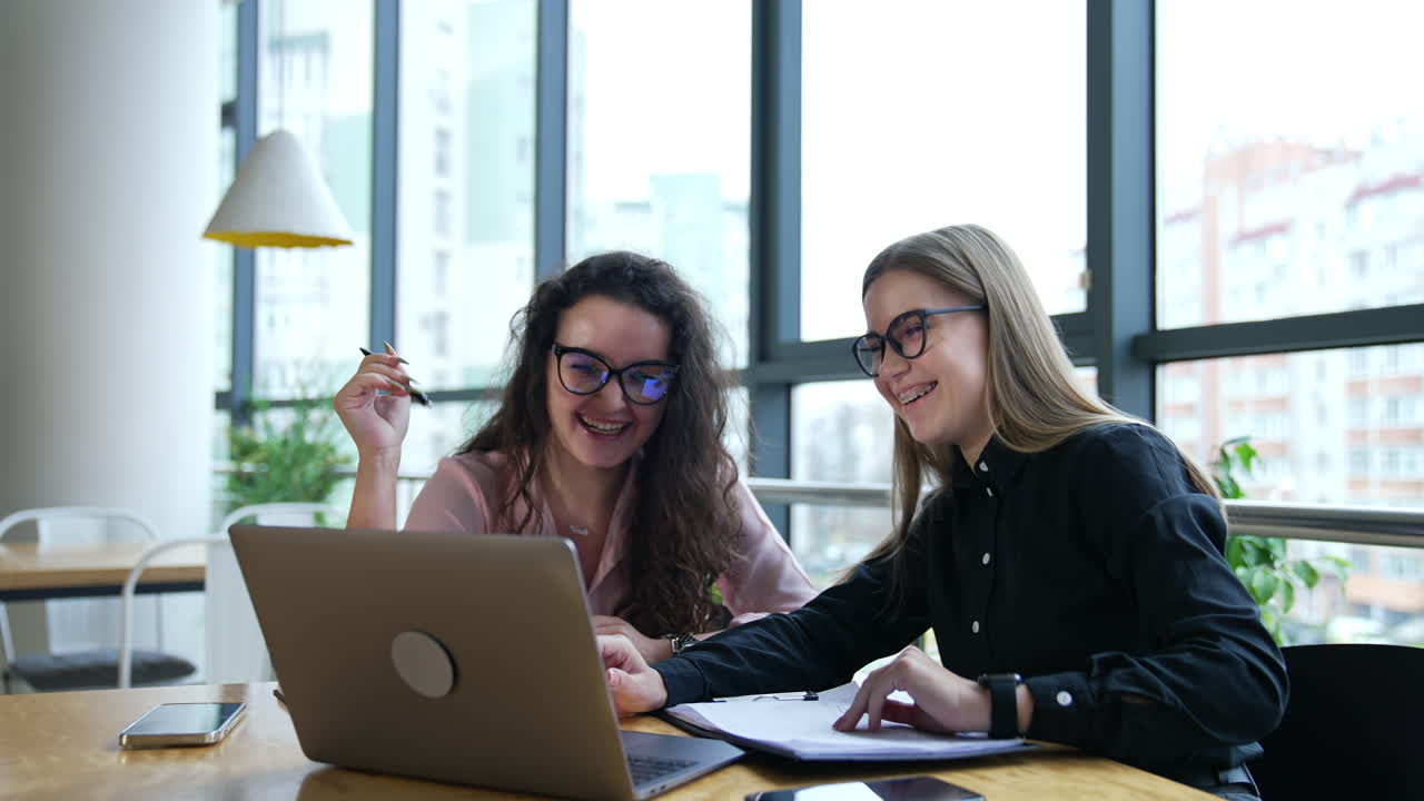 Women look at the laptop and laugh cheerfully. Two business ladies work at the computer in big light office. Businesswomen wearing glasses at work.