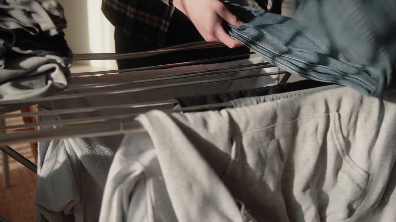 Young Male collecting and folding dry clean Clothes from Dryer At Home in sunlit modern living room grey t-shirt and blue jeans close up 4K