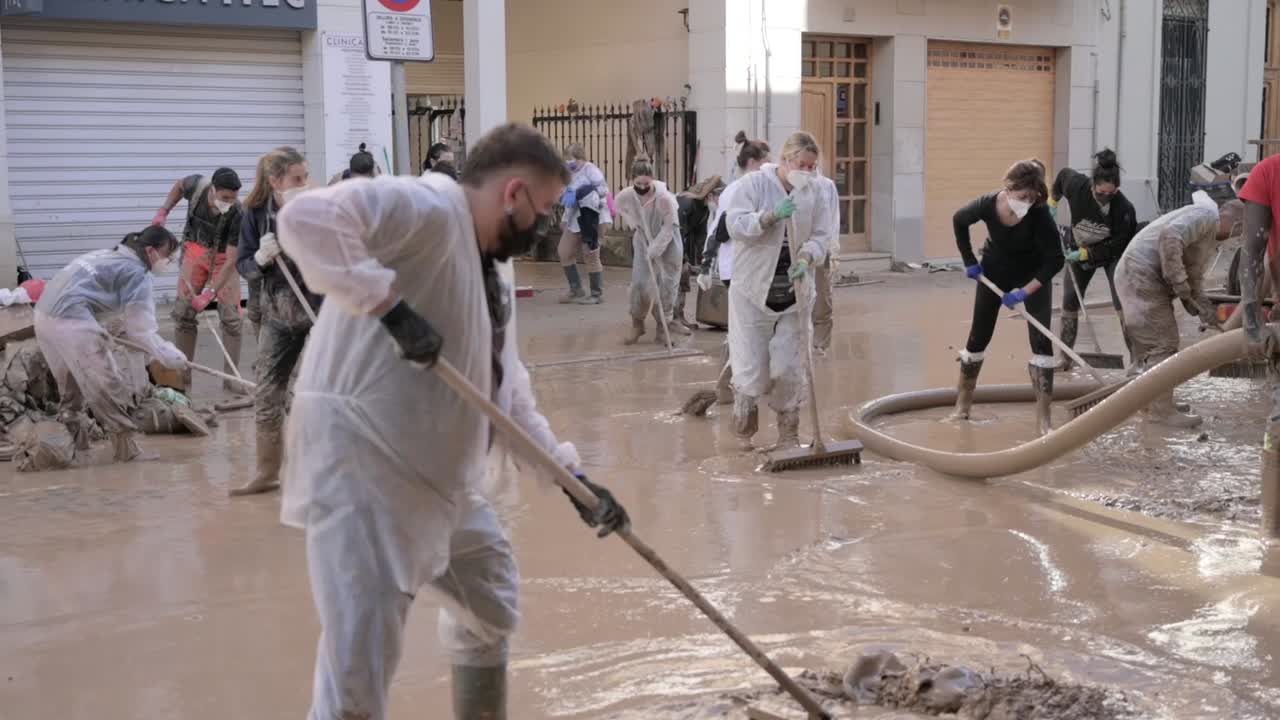 Community volunteers clean a flood-damaged street covered in mud. Wearing masks and protective gear, they show teamwork and resilience, supporting recovery efforts after the disaster