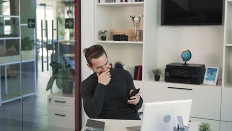 The-cheerful-young-man-is-sitting-at-a-desk-with-laptop-in-the-office