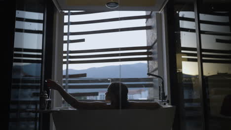 slow motion shot of a female lying in a bath looking out the window with a cocktail