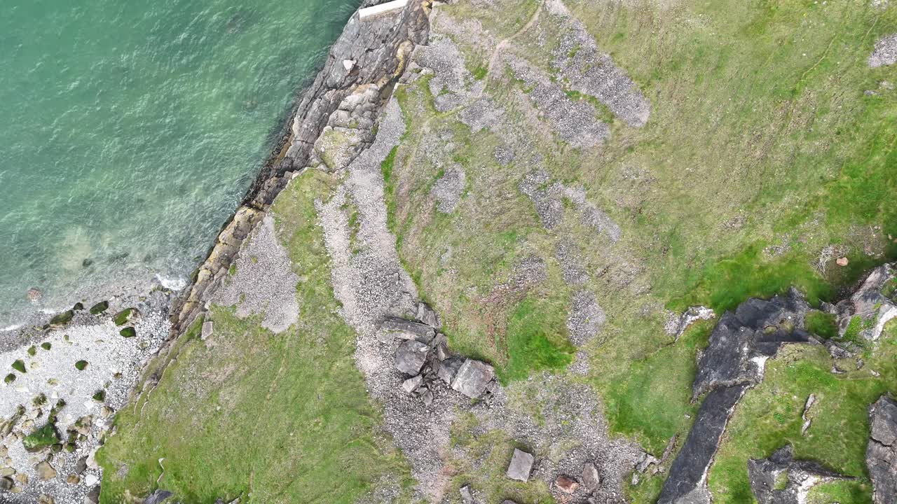 Drone video captures an aerial view of Wales’ rugged coastline, showcasing dramatic cliffs, lush greenery, and textured rock formations. Overcast lighting enhances the moody, natural scenery