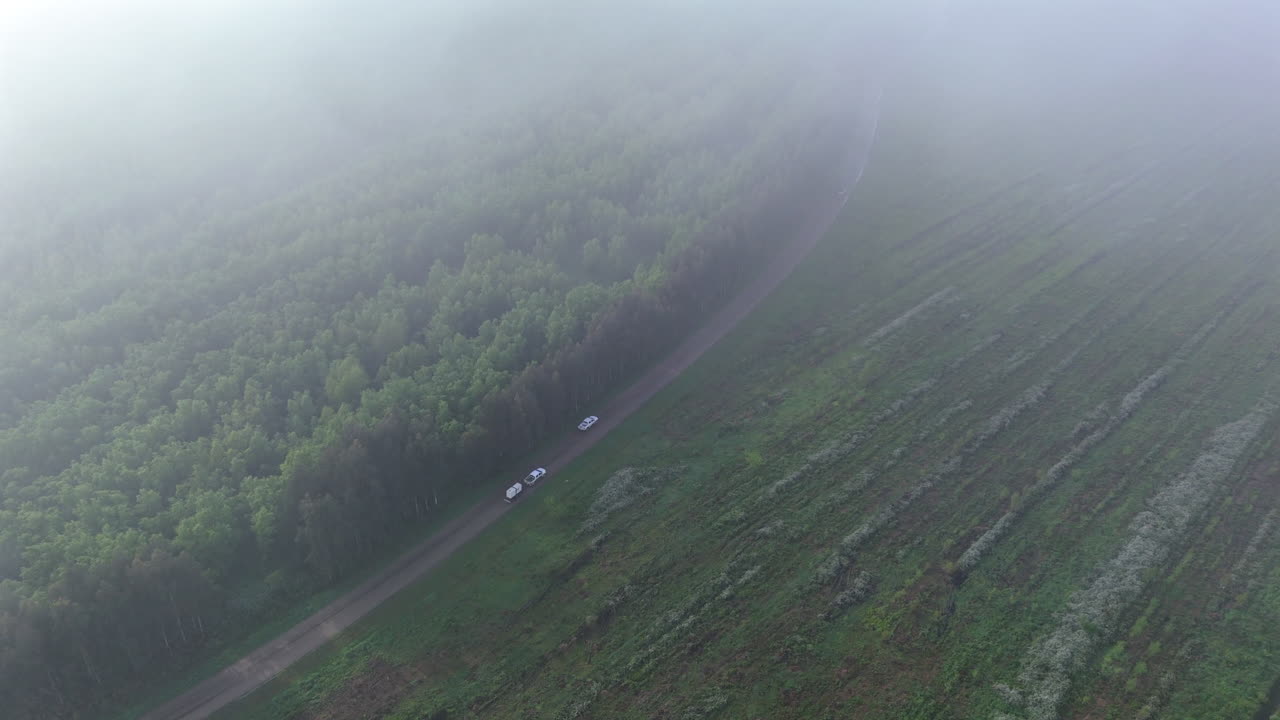Watch as drone soars through low clouds, capturing a car overtaking another with trailer on a rustic road. Surrounded by fields and forests, the scene offers a dynamic view of nature and movement.