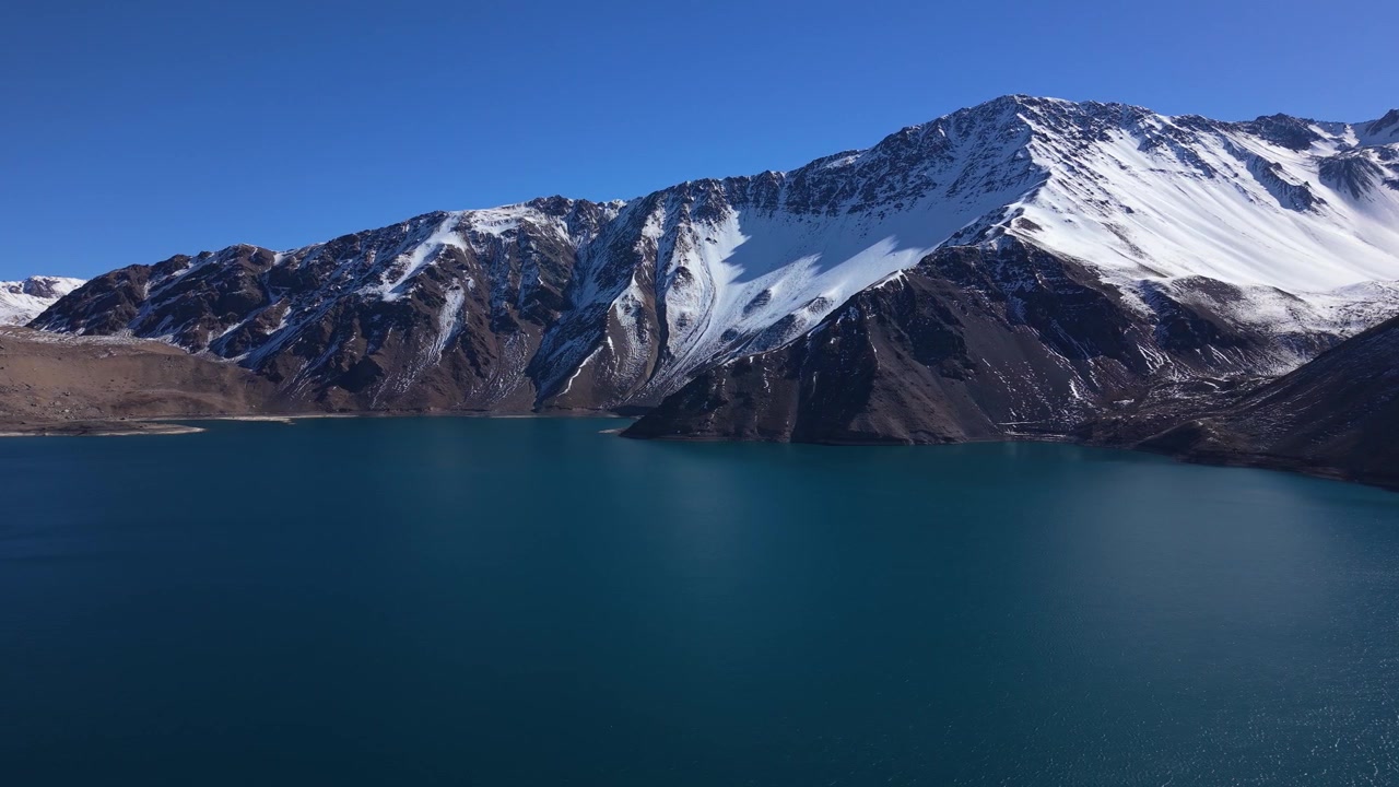 Lenta panoramica su acque glaciali di un blu profondo che rivela cime andine pesantemente innevate che si ergono ripide sotto un cielo limpido. Embalse del Yeso, Cile.