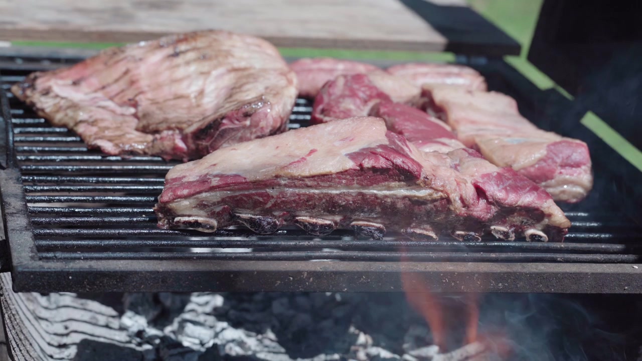 Close-up iga sapi, daging domba, dan sosis dipanggang di atas barbekyu api kayu dengan asap dan api. Pemandangan asado pedesaan di luar ruangan yang menampilkan budaya memasak tradisional Amerika Selatan