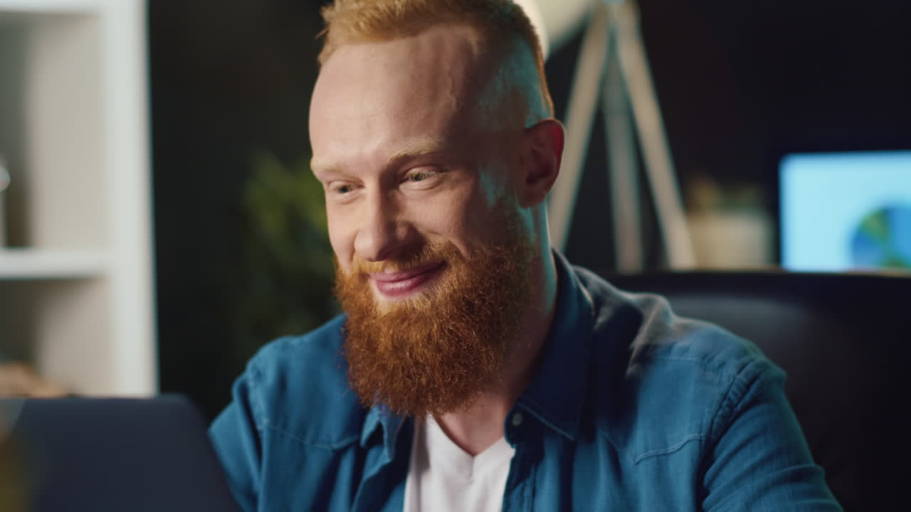 Cheerful man speaking on laptop at home. Redhead hipster working in office.