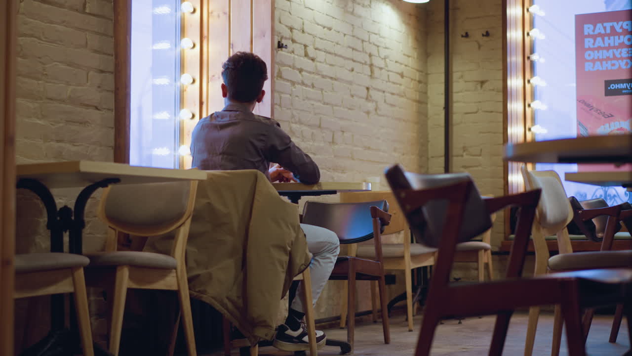 young man sitting alone at round table near window in empty cafe, coat draped over chair, back view, soft daylight, waiting, minimal interior, bag on bench, quiet contemplative mood