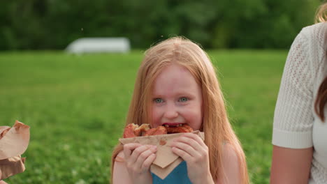 playful winking girl eating snack during family picnic in park, nature exploring child enjoying green grass, summer outdoor bonding moment with mother, carefree smile and joyful expression