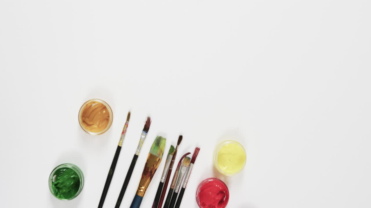 vista aérea de pinturas y pinceles sobre fondo blanco con espacio de copia, en cámara lenta