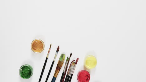 overhead view of paints and brushes on white background with copy space, in slow motion
