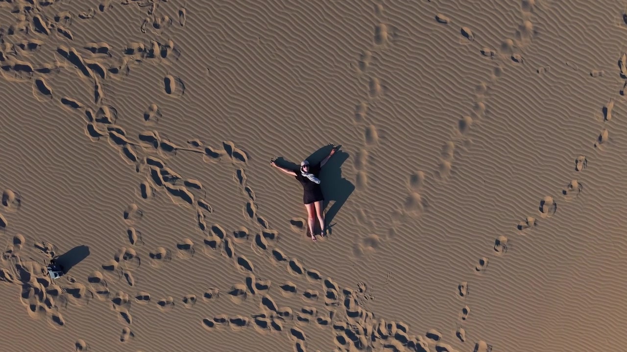Ripresa verticale con drone che si avvicina a una donna sdraiata sulla sabbia dorata ad Ammothines a Lemno, evidenziando un paesaggio desertico minimale e tonalità naturali tenui.