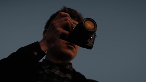 Young-caucasian-male-photographer-taking-picture-with-analog-photo-camera-during-sunset-against-the-blue-sky