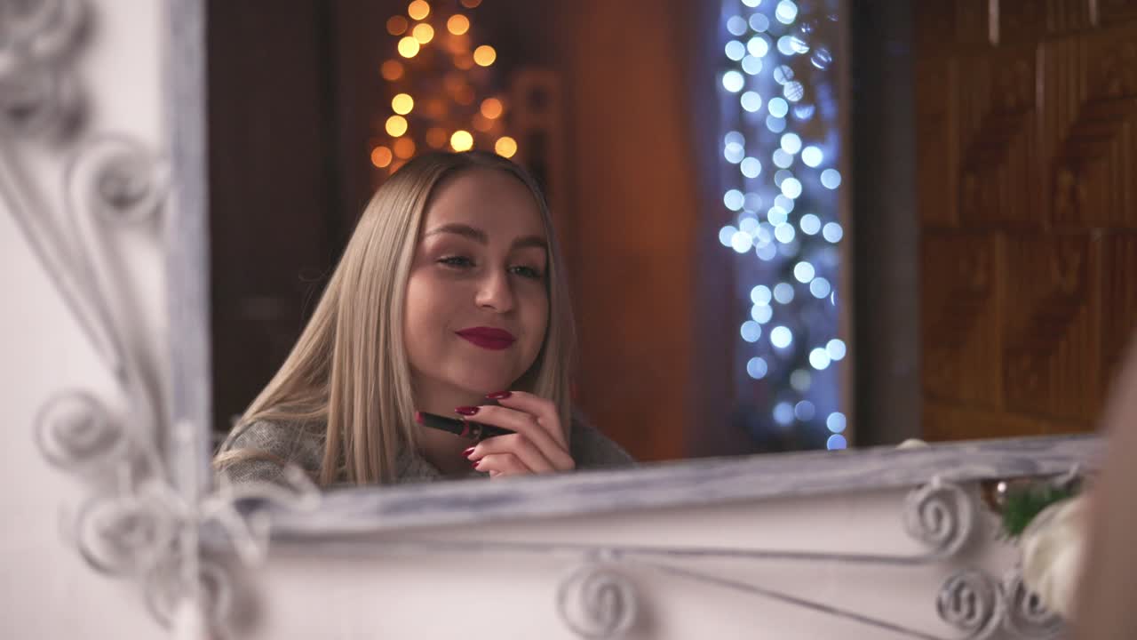 A young woman applies red lipstick while looking in the mirror, surrounded by warm and cool holiday lights. The soft glow and festive atmosphere create a cozy and elegant winter mood