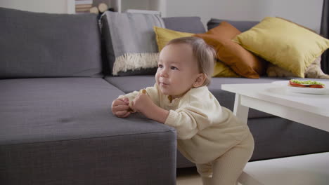 cute little girl standing next to sofa at living room and eating bread 1