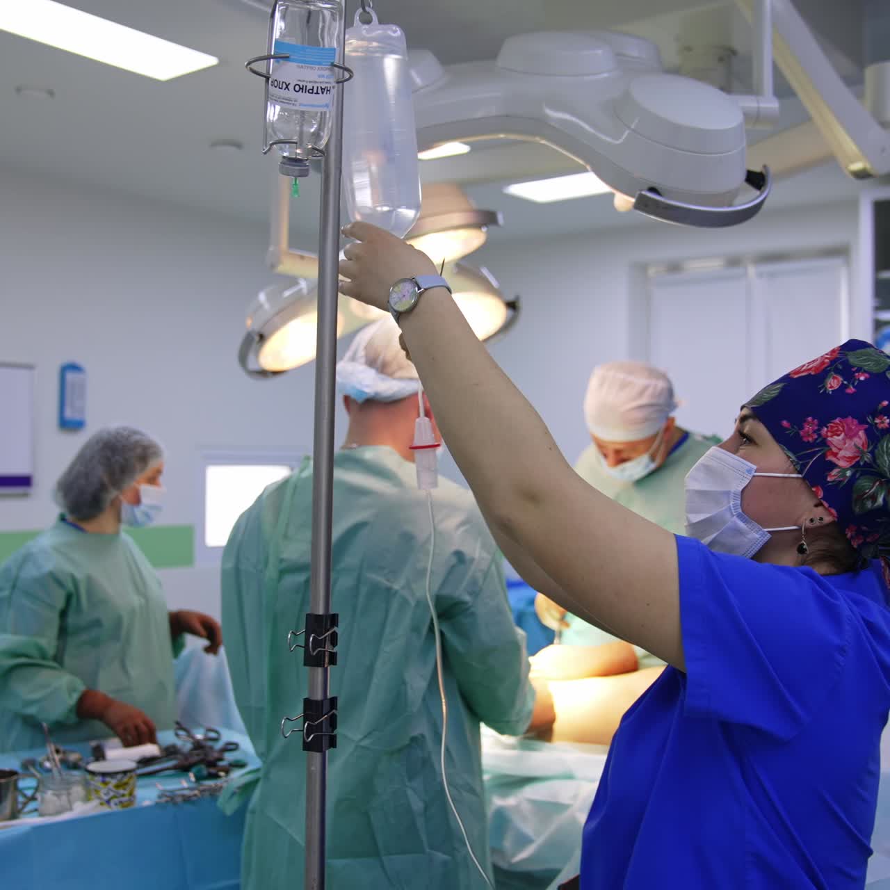 Nurse in mask and flowery cap attaching the bottle of medicines to the drop counter. Group of surgeons perform operational procedure at backdrop