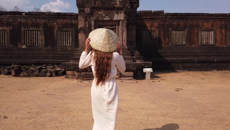 young beautiful woman in long white dress and vietnamese hat going to palace in wat phou ruined khmer hindu temple complex. champassak, laos, asia. sunny. ancient culture religious architecture. slow