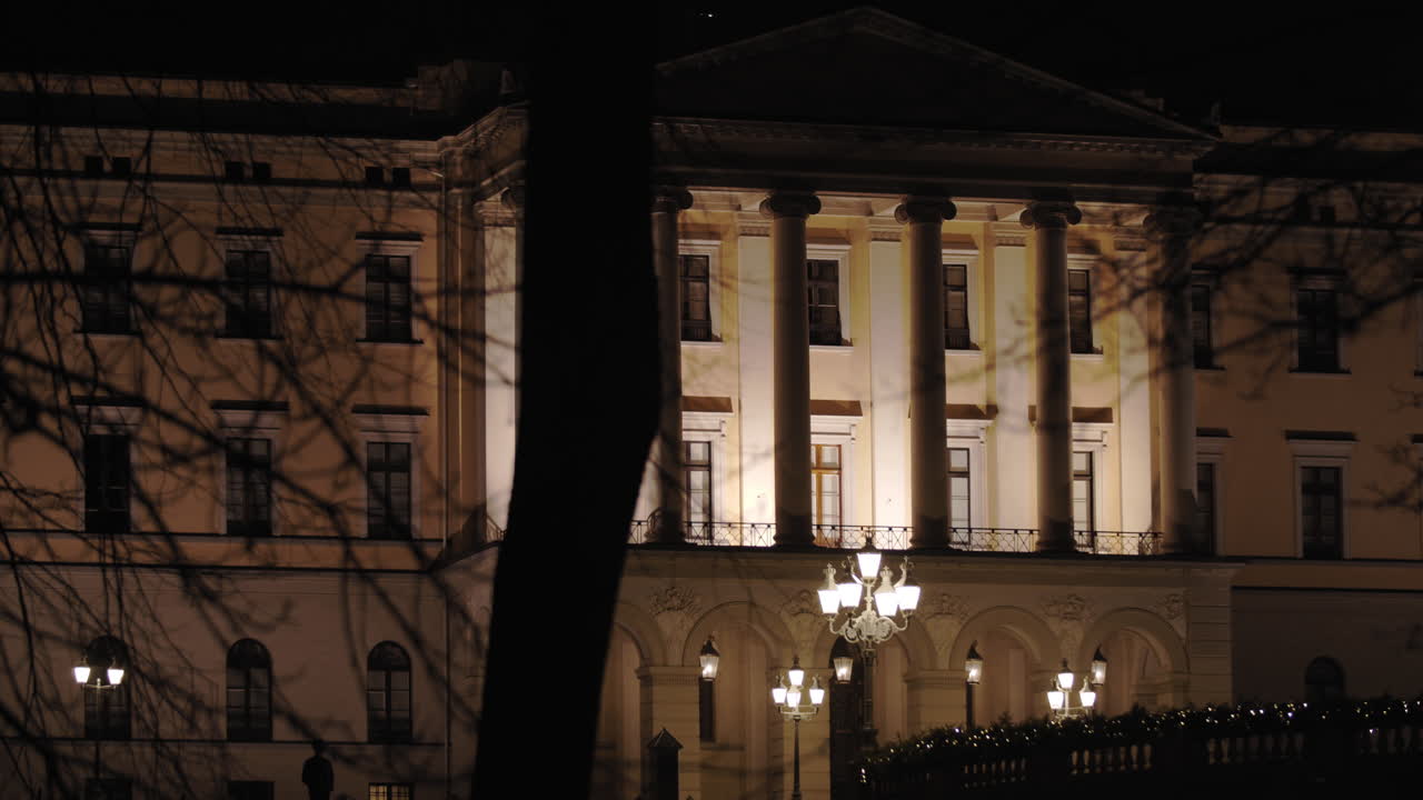 Slow motion wide 4K shot with parallax motion of the illuminated front of the Norwegian Royal Palace on top of Karl Johan street, with trees in foreground, at night in Oslo Norway